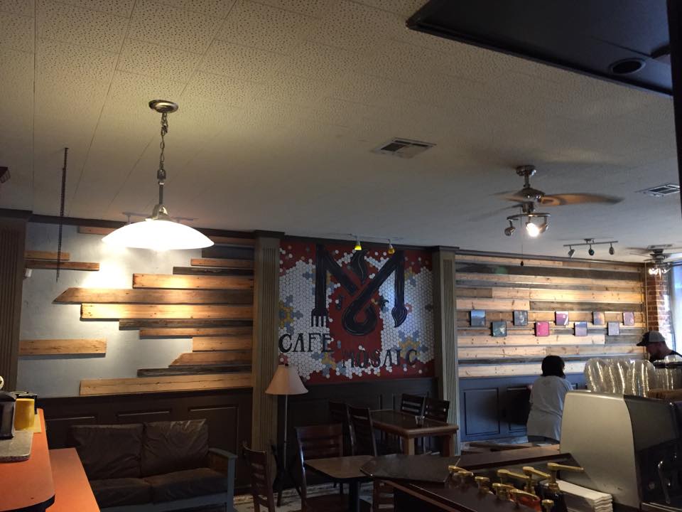 The warm interior of Cafe Mosaic — reclaimed-wood shelving, a hanging pendant lamp, and the tile mosaic mural on the back wall.