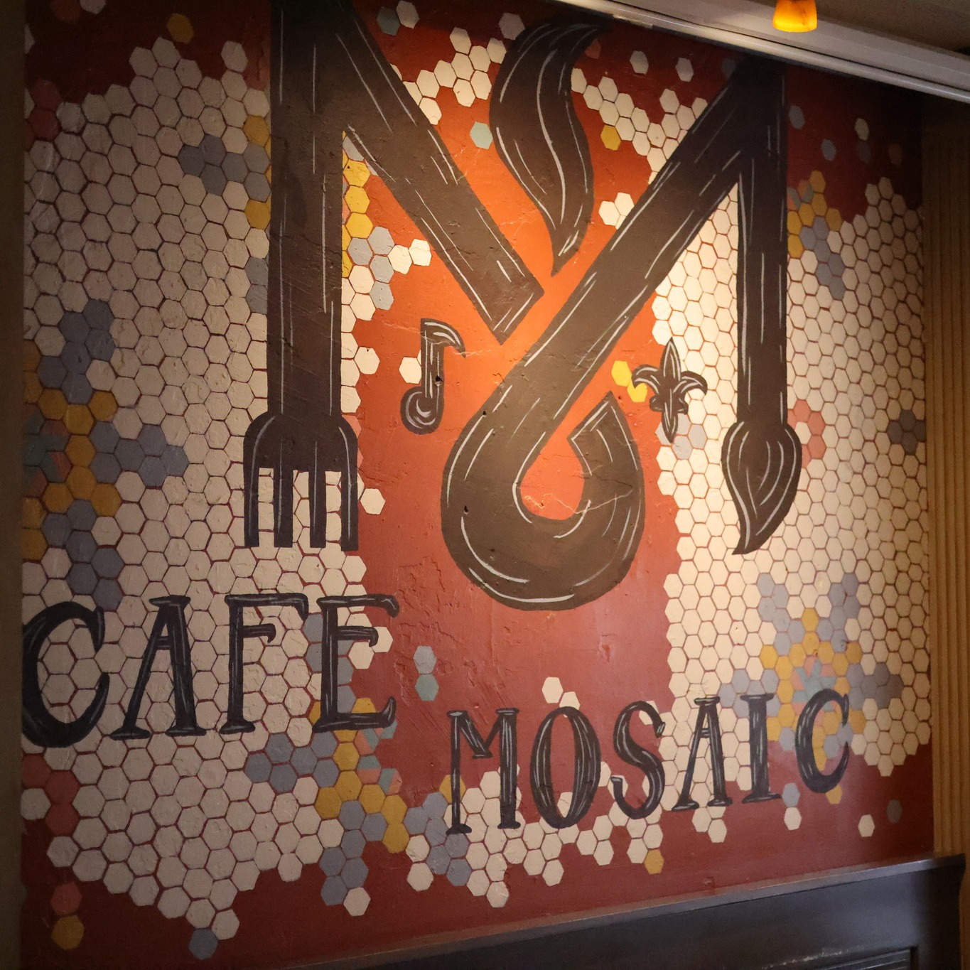 The hand-painted Cafe Mosaic mural — a fork, paintbrush, music note and coffee steam forming an 'M' over a honeycomb of mosaic tiles.