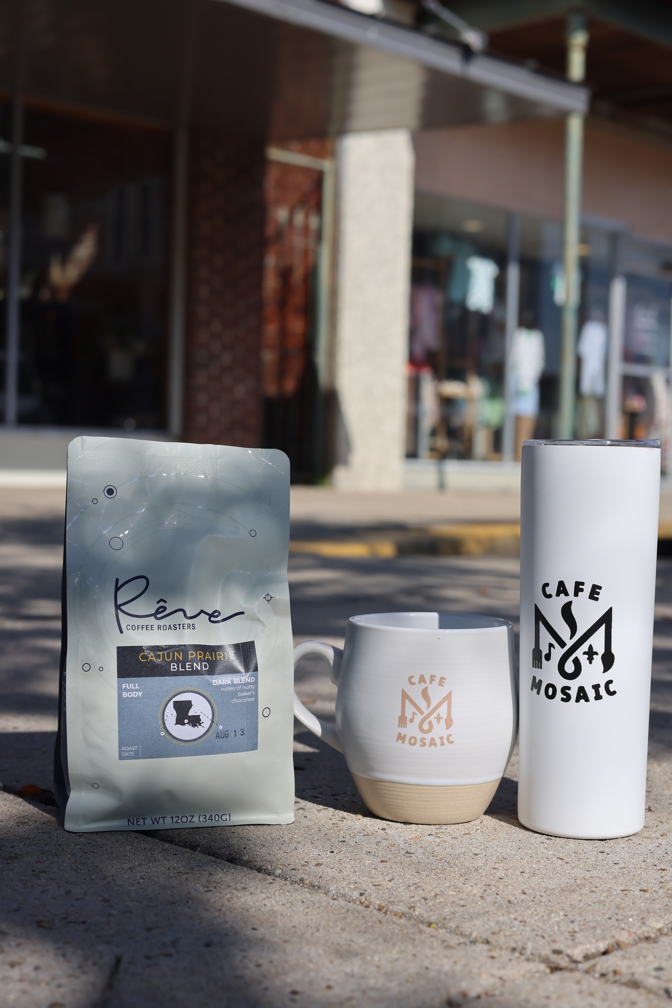 A bag of Rêve Cajun Prairie Blend with a Cafe Mosaic mug and travel tumbler outside the storefront.
